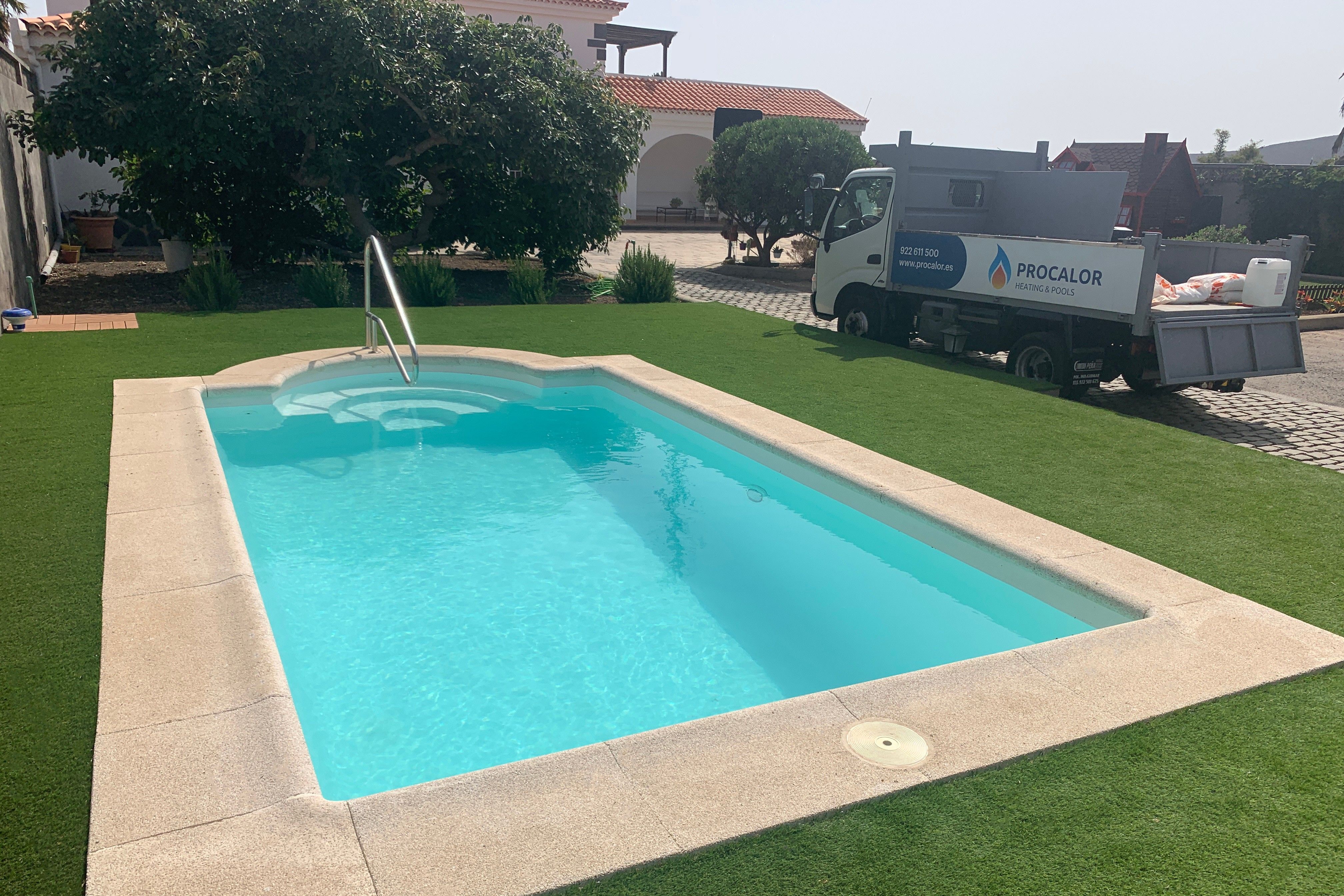 Piscina compacta recién instalada con pasamanos y borde de piedra en un jardín de césped, con el camión de PROCALOR al fondo
